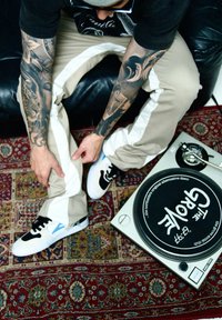 Čierno-biele tenisky s modrými detailmi, béžové nohavice s bielymi pruhmi a komplikované tetovania na rukách, umiestnené na vzorovanom koberci vedľa gramofónu.