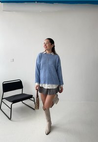 Svetle modrý pletený svetr, bílá košile s límečkem, vzorovaná šedá sukně, béžové vysoké boty až po kolena a malá taška, s černou židlí v pozadí.