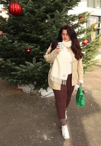 Giacca beige con sciarpa bianca frangiata, leggings marroni e borsa verde. Tiene in mano una tazza di caffè a righe, in piedi accanto a un albero sempreverde decorato.