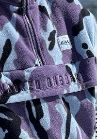 Chaqueta de forro polar con estampado de camuflaje en púrpura, azul claro y negro, con cierre de cremallera y una etiqueta de parche que presenta texto en relieve "ON BOARD" en el bolsillo delantero.
