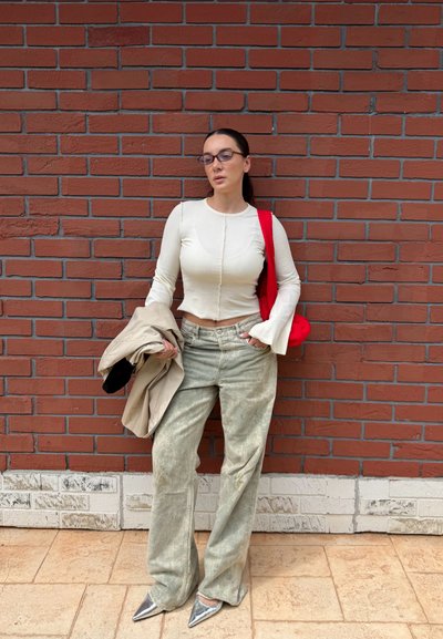 Mujer con gafas que lleva una camisa blanca de manga larga, pantalones grises sueltos, zapatos plateados, sosteniendo un abrigo beige, de pie contra una pared de ladrillos rojos.