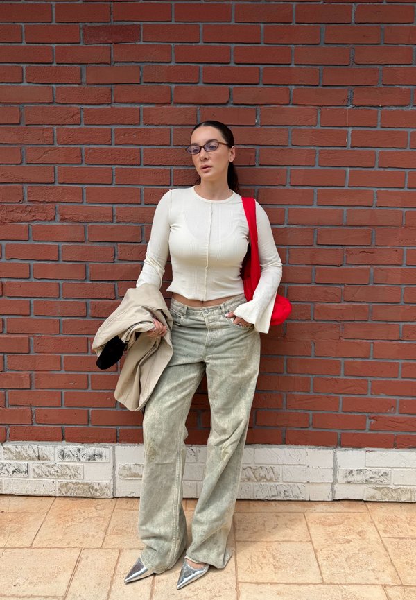 Femme avec des lunettes portant une chemise blanche à manches longues, un pantalon gris ample, des chaussures argentées, tenant un manteau beige, debout contre un mur en briques rouges.