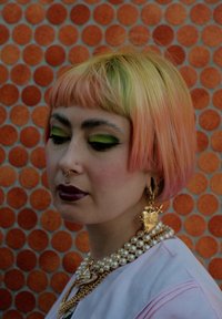 Krótko różowe włosy z zielonymi końcówkami, wyraziste zielone cienie do powiek i ciemna pomadka. Perły i złota biżuteria stanowią dodatki na tle w pomarańczowe kropki.