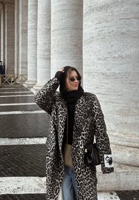 Femme portant un manteau à imprimé léopard, capuche noire et gants, lunettes de soleil, debout près de grandes colonnes en pierre blanche sur un sol pavé.
