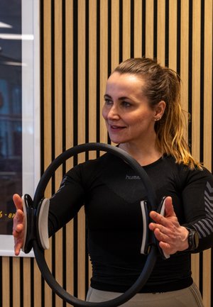 Mujer con ropa de entrenamiento negra sosteniendo y estirando un aro de pilates con un fondo de pared de listones de madera.