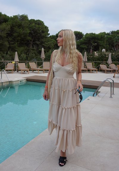 Vestido maxi en beige, con capas en volantes y un corsé ajustado, y tirantes ajustables. Combinado con sandalias negras de tiras y sosteniendo unas gafas de sol.