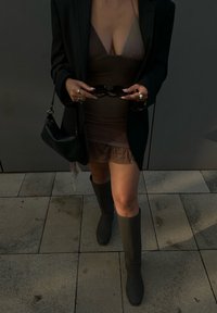 Vestito slip marrone con orlo in pizzo, indossato sotto un blazer nero a righe. Abbinato a stivali grigi sopra il ginocchio e con occhiali da sole neri in mano. Borsa nera.