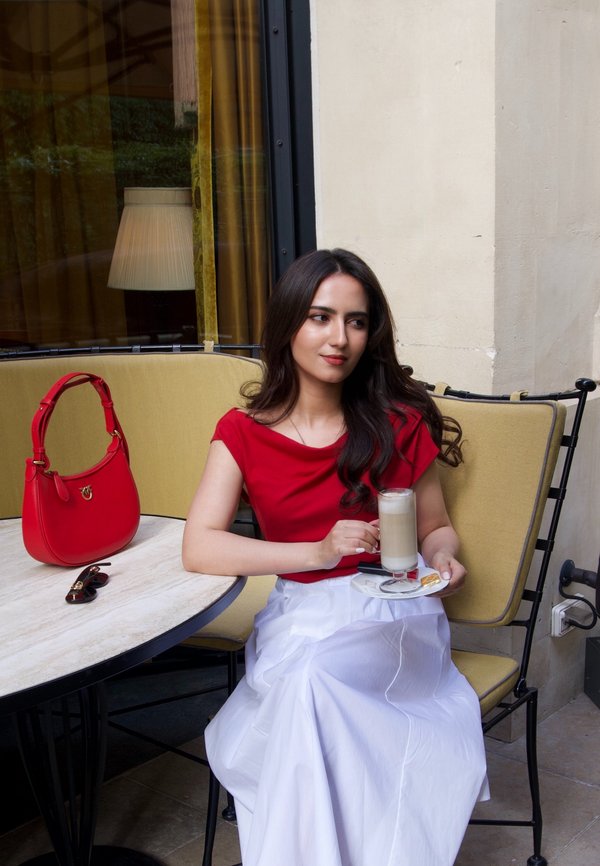 Femme en haut rouge et jupe blanche assise sur un banc rembourré jaune tenant un verre de latte à une table de café avec un sac à main rouge et des lunettes de soleil.