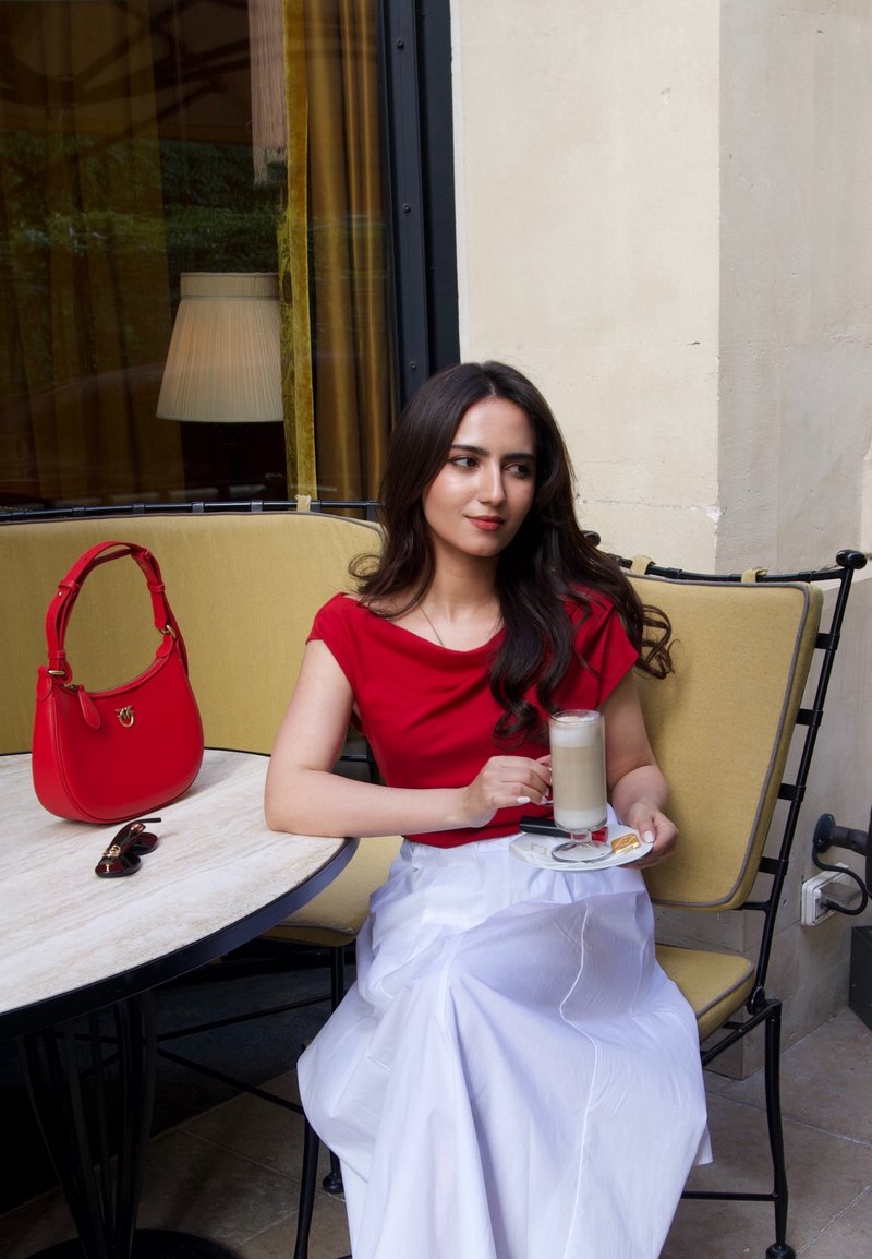 Woman in red top and white skirt sitting on a yellow cushioned bench holding a glass of latte at a cafe table with a red handbag and sunglasses.