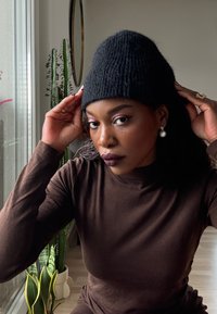 Bonnet en tricot noir porté par une personne aux cheveux foncés. En arrière-plan, des plantes en pot et un miroir circulaire. Haut marron à manches longues.