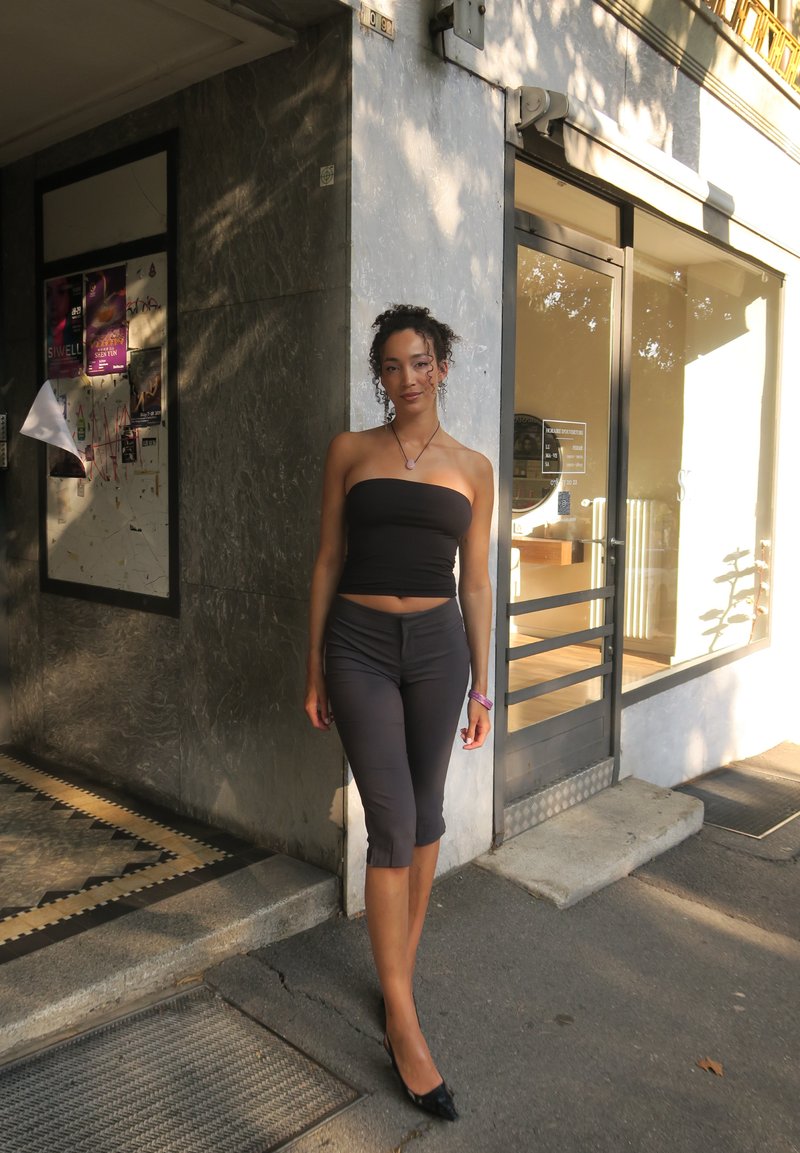 Jeune femme en top bandeau noir et pantalon court, debout sur le trottoir devant l'entrée d'un bâtiment en pleine journée.