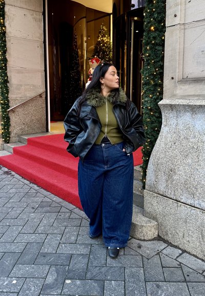 Chaqueta de cuero negra con cuello de piel sintética verde, suéter verde acanalado y vaqueros azules de pierna ancha. Escaleras conducen a una entrada festiva.