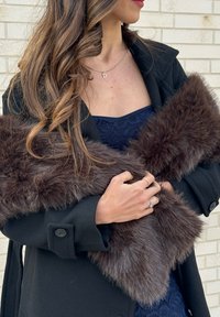 Femme aux longs cheveux bouclés portant un manteau noir et une robe en dentelle bleu marine, tenant un étole en fausse fourrure marron contre un mur en briques claires.