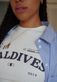Valkoinen puuvillapaita, jossa on tummansininen teksti "MALDIVES 2014", joka on sinivalkoisten raidallisten napitettu paidan alla. Yksinkertainen pyöreä kultaketju.