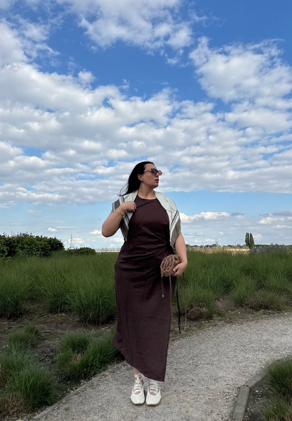 Femme en robe sombre et chaussures blanches marchant sur un chemin à côté de buissons verts sous un ciel bleu avec des nuages épars, tenant un petit sac.