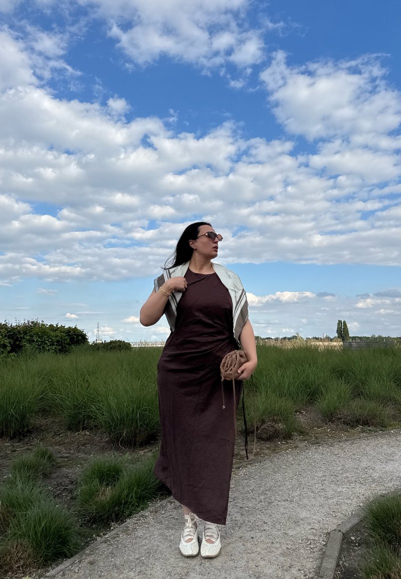 Woman in dark dress and white shoes walking on a path beside green bushes under a blue sky with scattered clouds, holding a small bag.