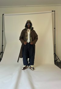Mann mit Sonnenbrille, Camouflage-Hoodie unter einem braunen Mantel, weißes Shirt, weite dunkle Jeans und gemusterte Stiefel, der vor einem weißen Hintergrund steht.