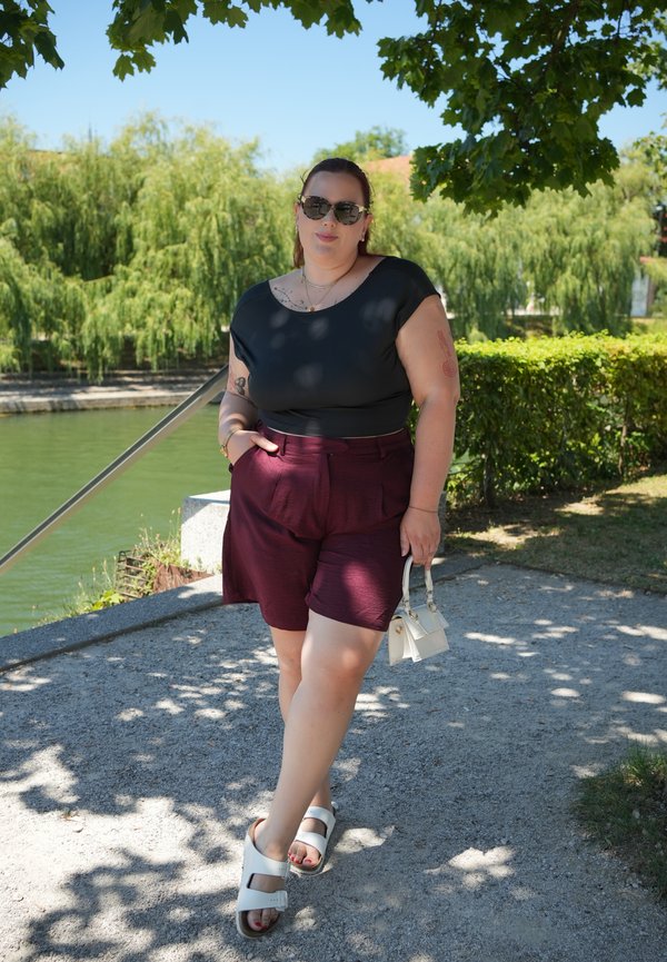 Femme portant des lunettes de soleil, un haut noir, un short bordeaux et des sandales blanches, debout sur un chemin au bord de l'eau avec des arbres et des buissons en arrière-plan.