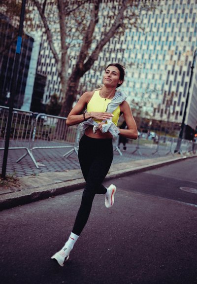 Joven mujer corriendo en la calle de la ciudad con un sujetador deportivo amarillo, leggings negros, zapatillas blancas para correr, sosteniendo una chaqueta gris con edificios y barreras cercanas.
