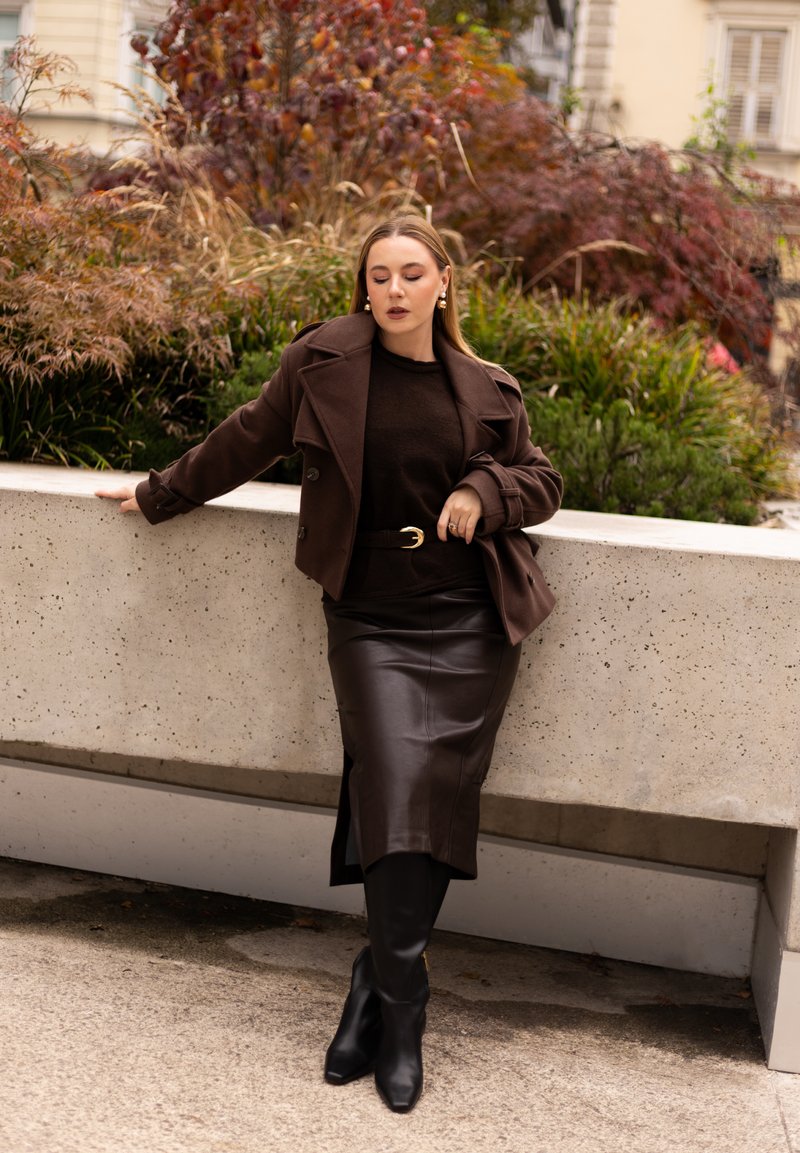 Manteau en laine brun sur un pull en tricot brun, associé à une jupe crayon en cuir brun et des bottes noires jusqu'aux genoux; cadre extérieur avec des plantes.