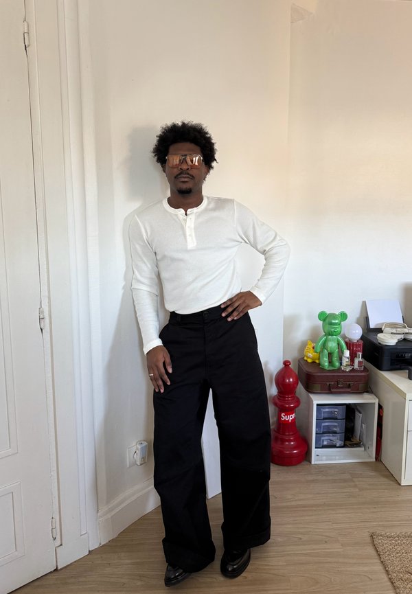 Man met krullend haar en zonnebril, gekleed in een wit henley-shirt en zwarte wijde broek, staand binnen bij planken met decoratieve voorwerpen.