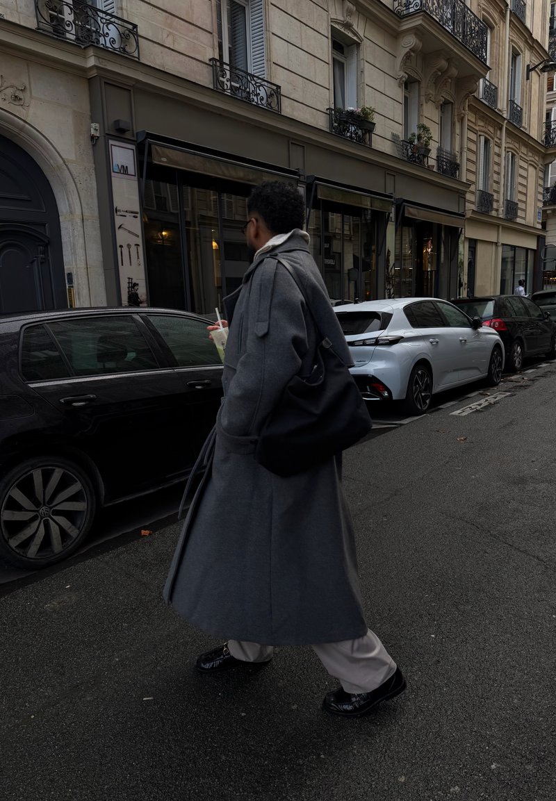 Personne en manteau gris long marchant dans une rue de la ville en tenant une boisson, avec des voitures garées et des immeubles à plusieurs étages en arrière-plan.