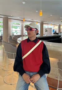 A person is wearing a red sweater over a black shirt, teamed with light blue jeans and red sunglasses. A beige cap finishes off the outfit.