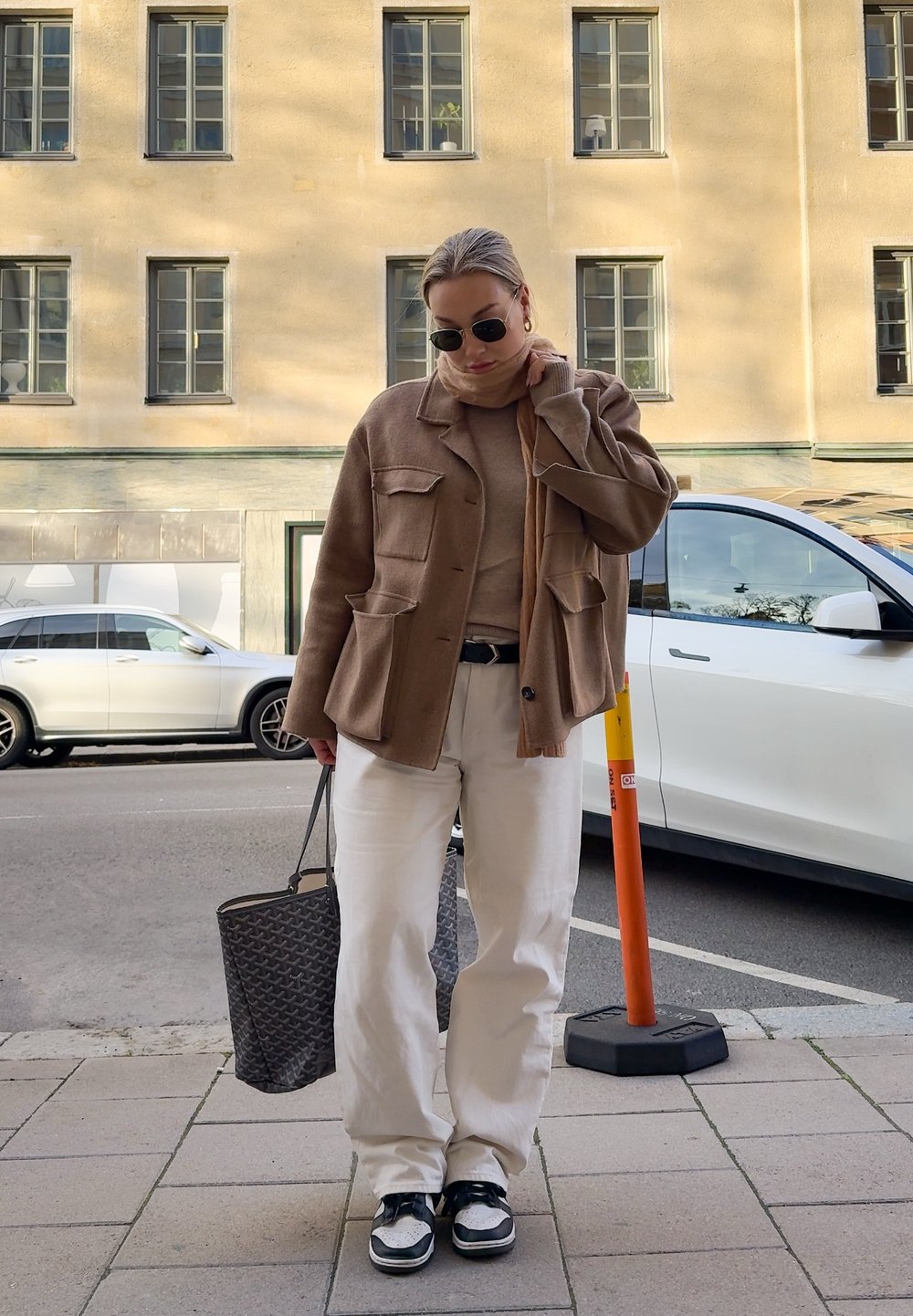 Braune Wollmischung Jacke, beiger Strickpullover, weiße weite Hose, schwarz-weiße Sneakers, strukturierte schwarze Tasche, Sonnenbrille, urbaner Hintergrund.