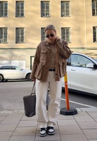 Braune Wollmischung Jacke, beiger Strickpullover, weiße weite Hose, schwarz-weiße Sneakers, strukturierte schwarze Tasche, Sonnenbrille, urbaner Hintergrund.
