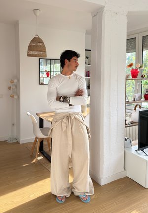Pantalones de pierna ancha beige con cintura anudada, camisa blanca de manga larga con canalé y chanclas azules. Suelo de madera natural y paredes blancas en una habitación luminosa.