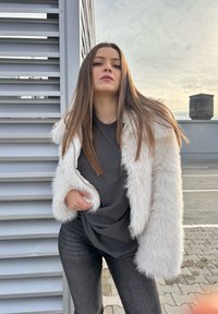 Bijela jakna od umjetnog krzna dugih rukava, sivi majica ispod i tamnosive uske traperice. Glatka tekstura s ležernim krojem.
