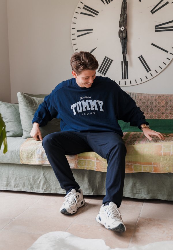 Sudadera de gran tamaño en color navy con el logo "TOMMY JEANS", combinada con pantalones negros y zapatillas blancas. Sentado en un sofá verde con un cojín estampado.