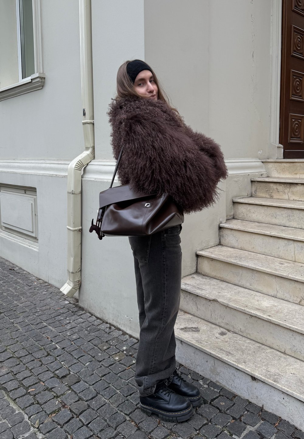 Manteau en fausse fourrure marron duveteux, jean denim foncé, chaussures compensées noires, et sac à main en cuir marron avec rabat et détail en métal.