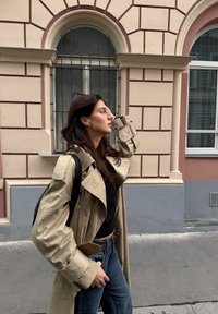 Bežový trenčkot s širokými klopami, černý top a modré džíny s vysokým pasem. Drží telefon a nosí černý batoh. Městské prostředí s architekturou.
