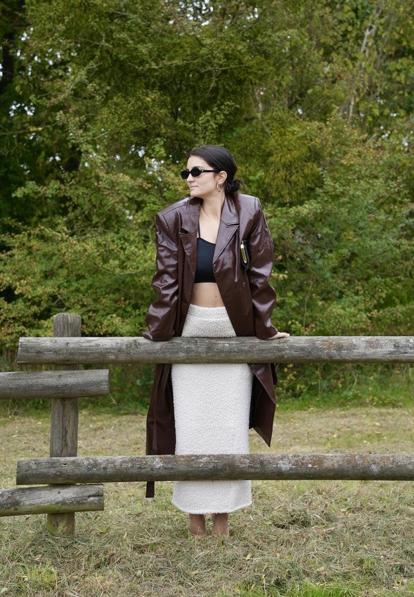 Manteau long en cuir marron, crop top noir, jupe blanche texturée et lunettes de soleil. Debout près d'une clôture en bois dans une zone herbeuse.