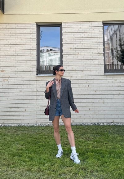 Mujer con gafas de sol que lleva un blazer gris, blusa rosa, pantalones cortos vaqueros, calcetines blancos y zapatillas está de pie sobre la hierba junto a un edificio de ladrillo claro con dos ventanas.