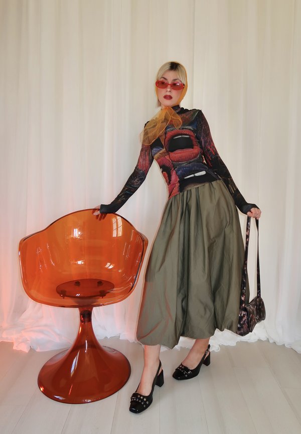 Nainen, jolla on punaiset aurinkolasit, pitkähihainen kuvioitu paita, oliivinvihreä hame, mustat korot, pitää laukkua, nojaten läpinäkyvään oranssiin tuoliin valkoisen verhon edessä.