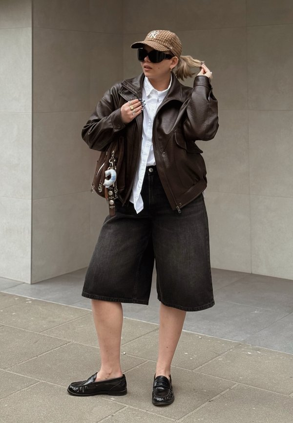 Osoba oblečená v hnedej koženej bunde, bielej košeli, čiernych širokých kraťasoch, čiernych mokasínach, károvanom klobúku a veľkých čiernych slnečných okuliaroch, drží si vlasy a tašku.