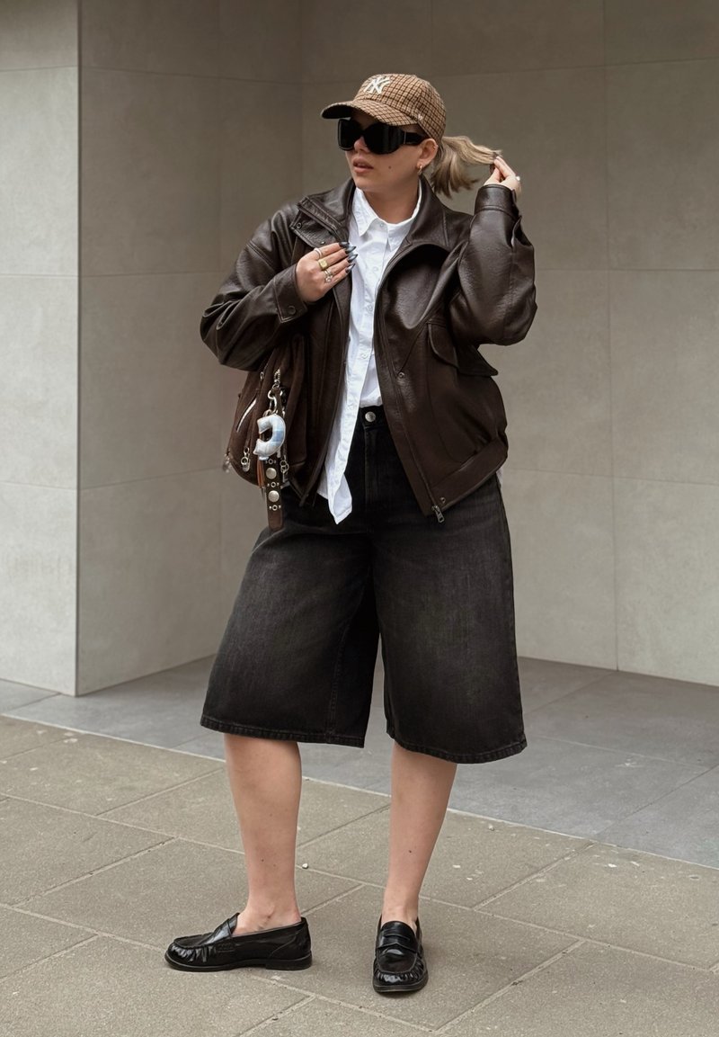 Άτομο με καφέ δερμάτινο μπουφάν, λευκό πουκάμισο, μαύρα φαρδιά σορτς, μαύρα μοκασίνια, καρό καπέλο και μεγάλα μαύρα γυαλιά ηλίου, κρατώντας τα μαλλιά και την τσάντα.