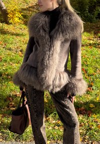 Jachetă maro cu blană la margini, cu textură moale și croială lejeră. Purtați-o peste un pulover negru cu guler înalt, asortată cu blugi gri cu model și o geantă maro.