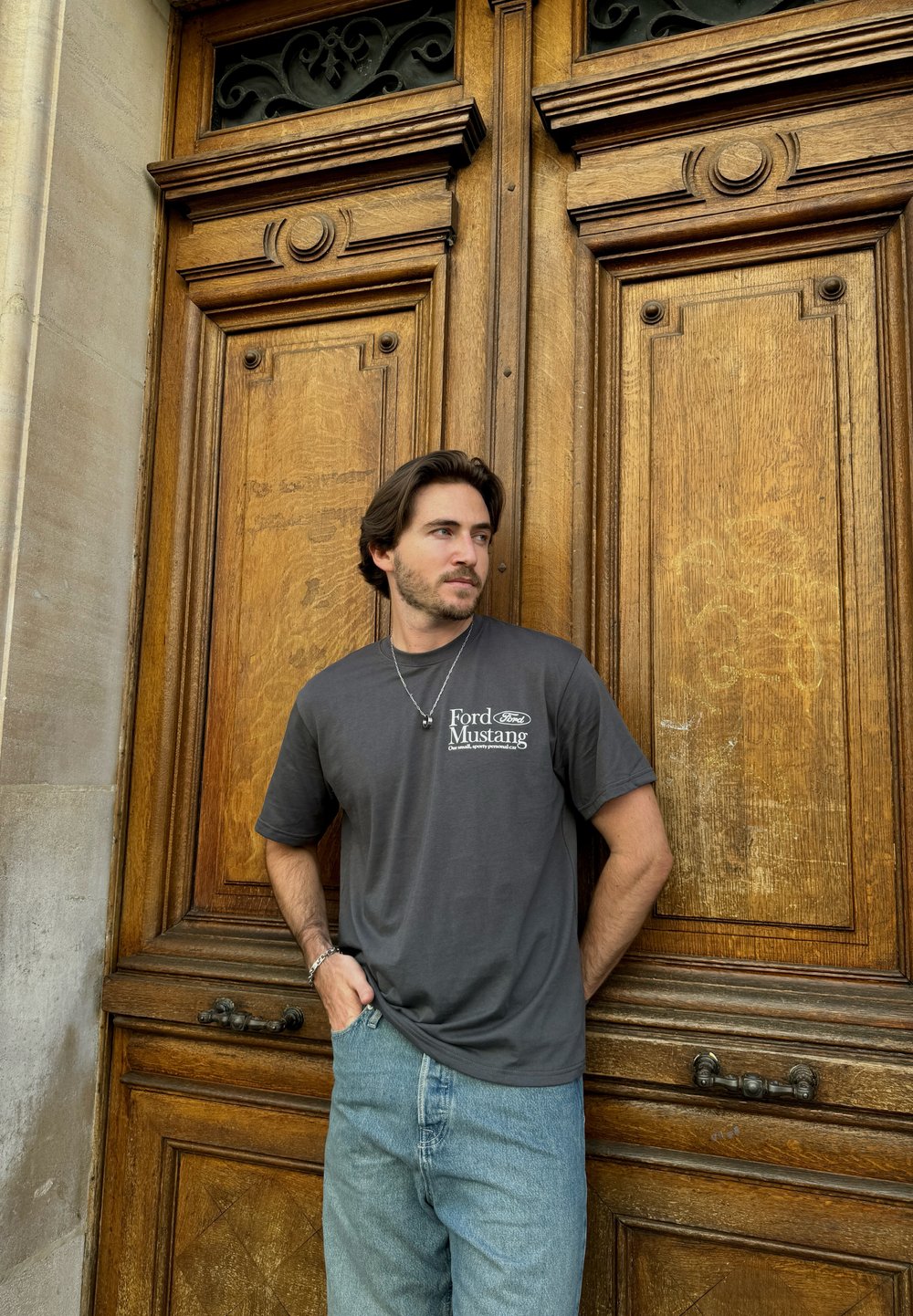 Γκρι μπλουζάκι με το λογότυπο "Ford Mustang" στην αριστερή πλευρά του στήθους, σε συνδυασμό με ανοιχτό μπλε τζιν παντελόνι. Φόντο με ξύλινες πόρτες.