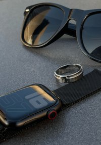 Črn pametni telefon z mrežnim pasom, črne sončna očala z debelimi okvirji in srebrn prstan z vgraviranim besedilom na sivi površini.