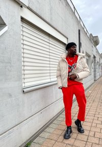 Puffer jacket in light beige with a high collar; red graphic t-shirt; red sweatpants; black chunky boots; standing against a concrete wall.