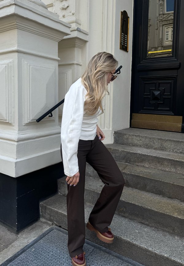 Femme aux longs cheveux blonds portant des lunettes de soleil, un haut blanc et un pantalon marron descendant des escaliers en pierre à côté d'une porte noire et d'une façade de bâtiment blanche.