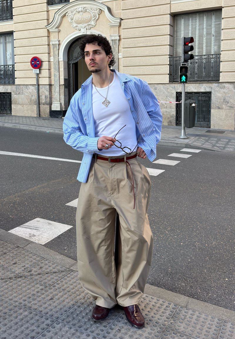 Un jeune homme aux cheveux bouclés se tient à un coin de rue, portant une chemise rayée bleue, un t-shirt blanc, un pantalon large beige, tenant des lunettes près du passage pour piétons.