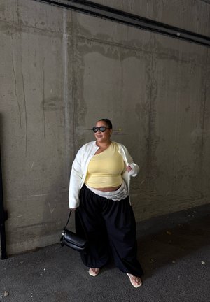Mujer con top amarillo, cárdigan blanco, pantalones anchos negros, gafas de sol y chanclas, sosteniendo un bolso negro, de pie contra una pared de hormigón.