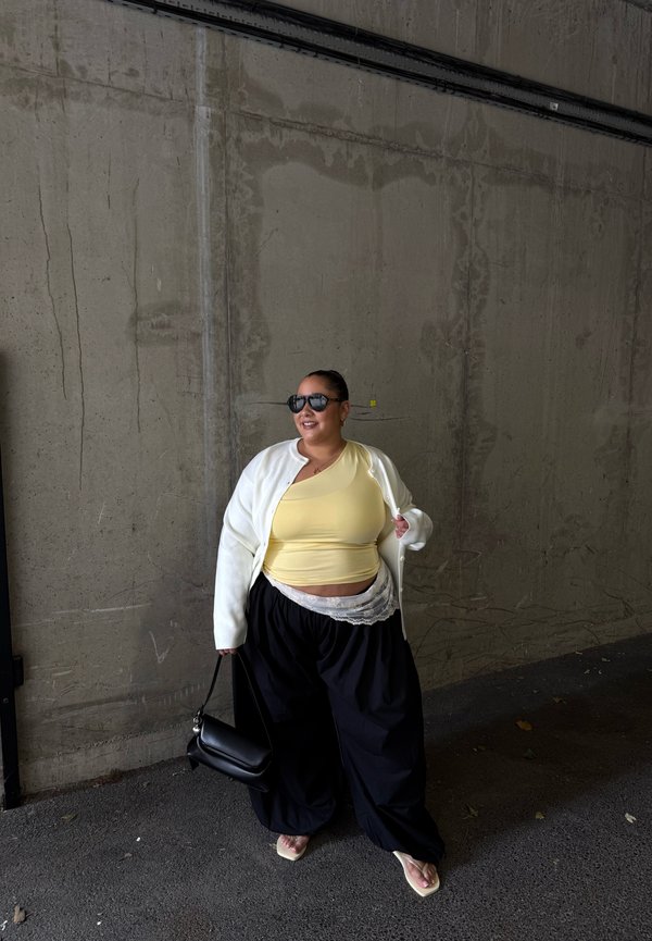 Femme portant un haut jaune, un cardigan blanc, un pantalon large noir, des lunettes de soleil et des tongs, tenant un sac à main noir, debout contre un mur en béton.