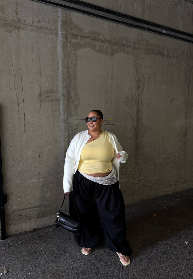 Femme portant un haut jaune, un cardigan blanc, un pantalon large noir, des lunettes de soleil et des tongs, tenant un sac à main noir, debout contre un mur en béton.