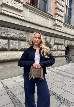 Joven mujer con cabello rubio largo está de pie en una acera de la ciudad vistiendo un cárdigan azul marino y pantalones, sosteniendo un bolso marrón con flecos.