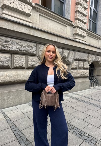Joven mujer con cabello rubio largo está de pie en una acera de la ciudad vistiendo un cárdigan azul marino y pantalones, sosteniendo un bolso marrón con flecos.